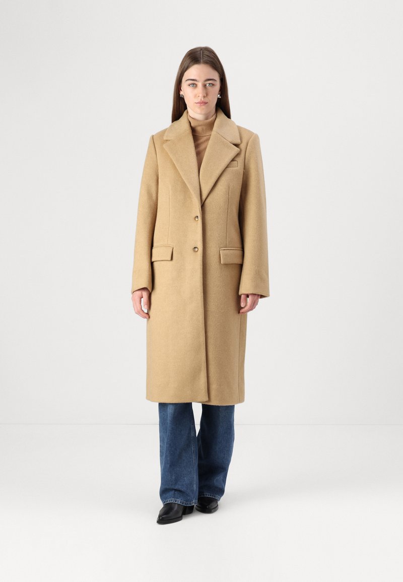 Lungo cappotto di lana beige con colletto a punta, due tasche frontali e chiusura con un solo bottone, abbinato a pantaloni in denim svasati.