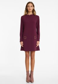 Woman standing wearing a long-sleeve plum dress with two gold buckle accents at the hips and light brown ankle boots.