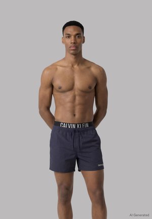 Uomo muscoloso in piedi contro uno sfondo grigio, che indossa pantaloncini da bagno Calvin Klein blu scuro con fascia in vita firmata e logo sulla gamba.