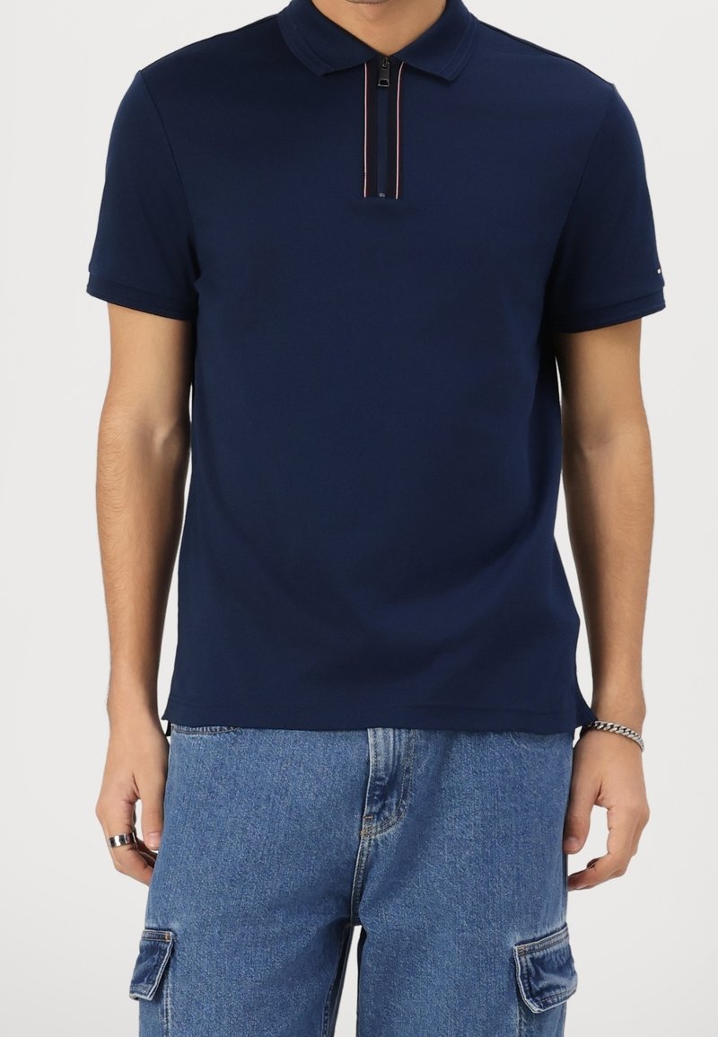 Polo shirt bleu marine avec un col zippé orné de rayures rouges et blanches. Fabriqué en tissu lisse, design ajusté, manches courtes.