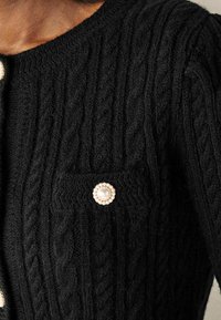 Czarny dzianinowy kardigan z wzorem w warkocz, wyposażony w guzik ozdobiony perłą oraz małą kieszonkę po lewej stronie.