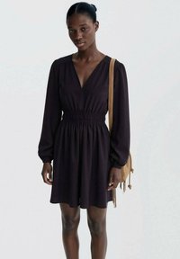 Temno vijolična obleka z izrezom v obliki V, dolgimi rokavi in nabranim pasom. Ujemajoča svetlo rjava torbica za čez ramo.