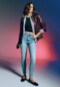 Rjav usnjen jopič čez zeleni kratki top, v kombinaciji s svetloblue ozkimi kavbojkami in črnimi balerinkami. Ozadje vsebuje gradient roza do modre.