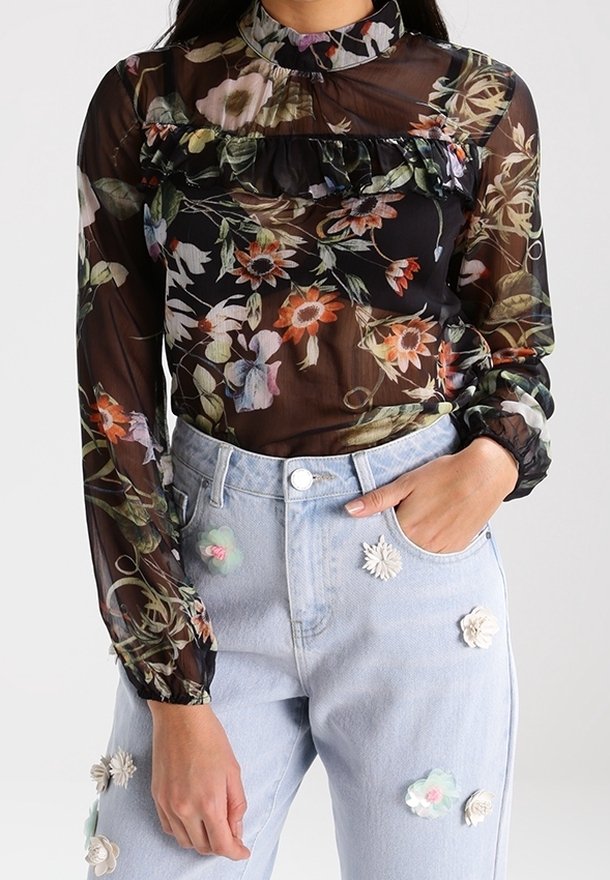 Blouse florale noire transparente avec des détails froncés au niveau de la poitrine, associée à un jean bleu clair orné d'appliqués floraux.