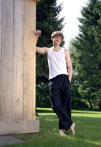 Vit linne, svarta löst sittande byxor, stående barfota mot en trästruktur med grönska i bakgrunden.