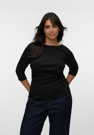 Kvinde med langt mørkt hår iført en sort top med trekvartærmer og mørkeblå jeans, stående med hænderne bag ryggen mod en hvid baggrund.