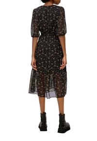 Svart blommig klänning med genomskinliga puffärmar, insvängd midja och lager på lager-kant. Bärs med svarta kraftiga stövlar. Bakgrunden är helt vit.