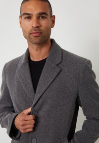 Grau Wolle Mantel mit Schalkragen, zwei Knöpfen und einem strukturierten schwarzen Strickpullover darunter. Glattes Material mit taillierter Passform.