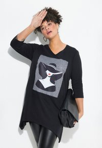 Tunic nera con scollo a V e maniche a tre quarti, decorata con una grafica bianco e nero di una donna. Indossata con leggings neri lucidi e una clutch.