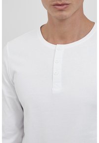 Hvit langermet henley skjorte laget av glat bomull, med rund hals, frontplakk med tre knapper og minimal sømdetalj.