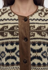 Veste en laine crème et marron à motifs, avec boutonnière sur le devant et col rond portée par une personne visible du menton à la poitrine.