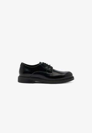 Zwarte leren veterschoenen met ronde neus en dikke zool, getoond in zijaanzicht op een witte achtergrond.