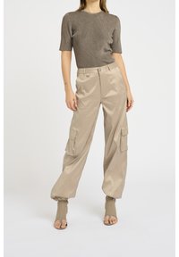 Beige cargo-housut, joissa on kiiltävä pinta, kahdella sivutaskulla ja ribatulla, lyhythihaisella tummalla paidalla. Kengät ovat avonaiset ja tiukat.