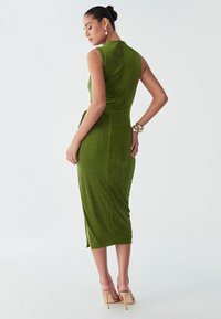 Model pozira u bezrukavnoj, zelenoj midi haljini s bočnim prorezom, ističući elegantne dodatke u minimalističkoj pozadini.
