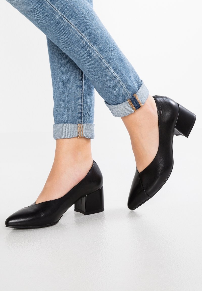 Femme portant un jean bleu retroussé et des chaussures à talons blocs en cuir noir, debout sur un sol blanc avec un pied légèrement levé.