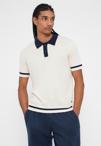 Krema polo majica z mornarsko barvo ovratnikom in mornarskimi črtami na rokavih; vsebuje dva gumba in tkanino z teksturo. Nosi se z mornarskimi hlačami.