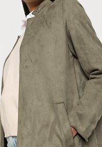 Persona che indossa un maglione beige chiaro e una giacca in suede verde oliva, con la mano in tasca, ripresa dalla testa ai fianchi, rivolta leggermente a sinistra.