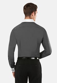 Camicia grigia a maniche lunghe con colletto bianco, dotata di polsini con bottoni e una texture liscia, indossata con pantaloni neri e una cintura nera.