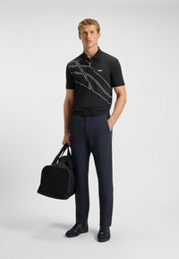 Czarna koszulka polo z białymi ukośnymi liniami, zestawiona z granatowymi spodniami. Mężczyzna trzyma czarną torbę sportową, nosi ciemne buty. Prosty, nowoczesny design.