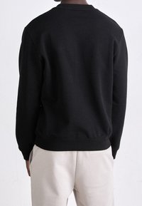 Sudadera negra con cuello redondo y puños acanalados, hecha de una tela suave. Presenta un ajuste holgado y está combinada con pantalones de color claro.