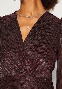 Femme portant une robe portefeuille plissée métallique violet profond et des colliers en argent superposés, dont un avec un pendentif blanc.