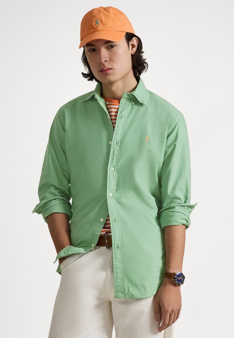 Grön skjorta med knappar och uppvikta ärmar, orange keps, randig T-shirt under, vita byxor och ett brunt bälte. Klocka på handleden.