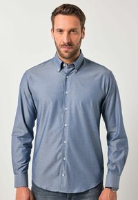 Uomo che indossa una camicia azzurra a maniche lunghe con motivo, con colletto abbottonato, in piedi davanti a uno sfondo chiaro e uniforme.