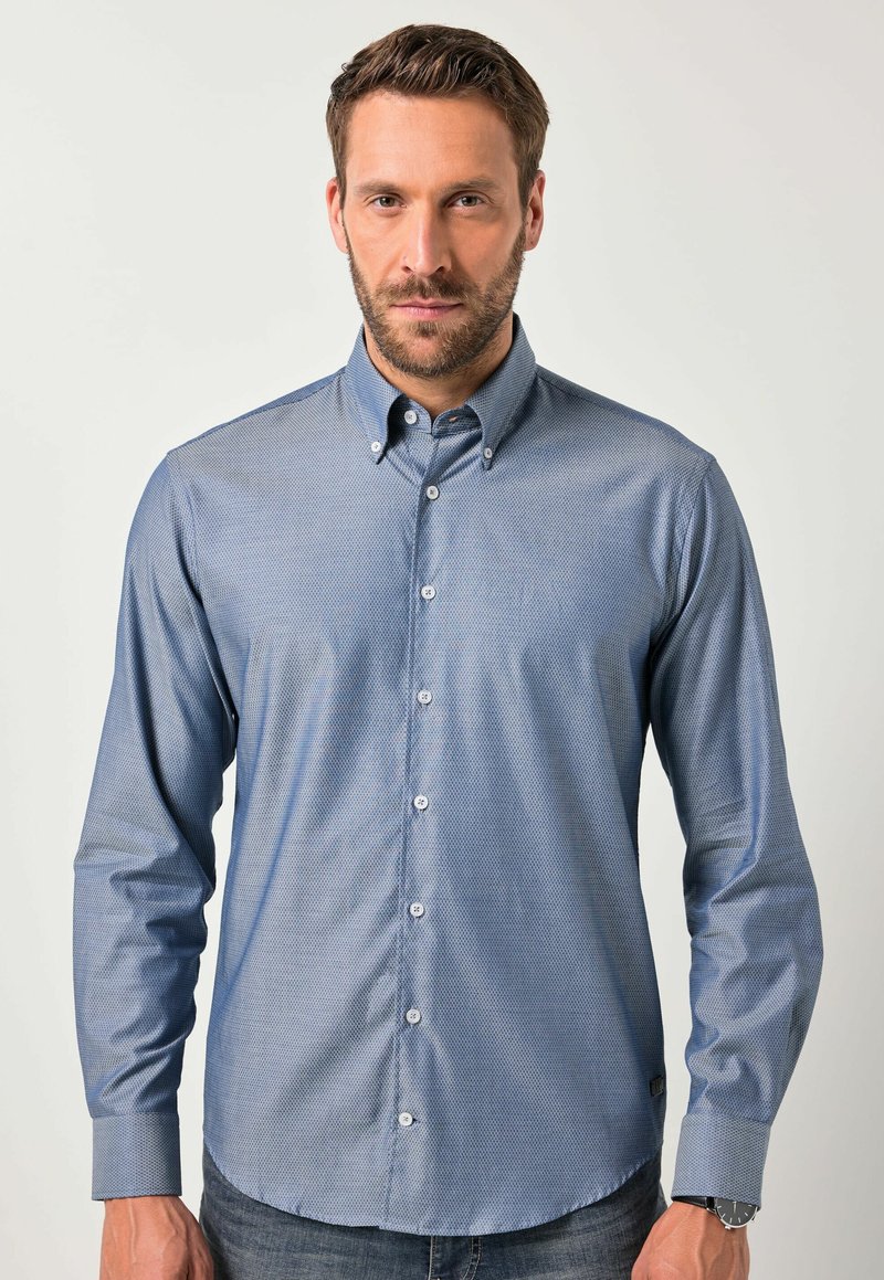 Uomo che indossa una camicia azzurra a maniche lunghe con motivo, con colletto abbottonato, in piedi davanti a uno sfondo chiaro e uniforme.