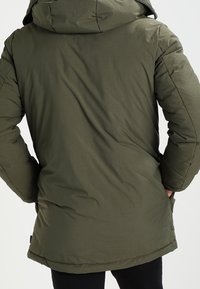 Ciemnozielona ocieplana kurtka z kapturem, o gładkiej fakturze i elastycznych mankietach. Widok od tyłu ukazujący prostą sylwetkę i ukryte kieszenie.