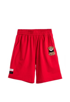 Joven con sudadera blanca con parche de osito, pantalones cortos rojos, calcetines blancos y botas marrones, de pie sobre fondo blanco.