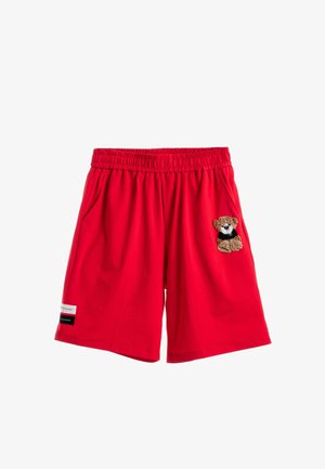 Joven con sudadera blanca con parche de osito, pantalones cortos rojos, calcetines blancos y botas marrones, de pie sobre fondo blanco.