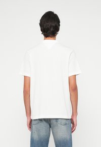 Mann som står med ryggen til iført en enkel hvit t-skjorte med korte ermer og falmet blå jeans, mot en ensfarget lys bakgrunn.