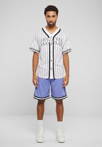 Hvidstribet trøje med "KANI" tekst, sorte accenter og sorte knapper, parret med lyseblå shorts med sort kant. Hvide sneakers.