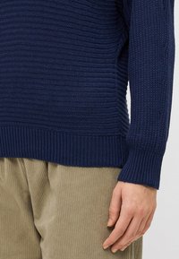 Marineblauwe gebreide trui met gestructureerde ribbel, voorzien van een brede manchette en zoom. Draagt over lichtbeige corduroy broek, hand zichtbaar bij de manchette.