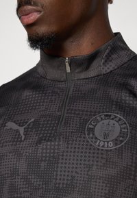 Fekete negyed cipzáras pulóver textúrált mintával. A mellkasán található a Puma logó és az FC St. Pauli embléma. Puha anyag, enyhe fényességgel.