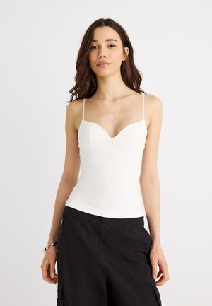 Γυναίκα με μακριά σκουρόχρωμα μαλλιά φοράει λευκή εφαρμοστή καλοκαιρινή μπλούζα με λεπτές τιράντες και μαύρο παντελόνι cargo, στέκεται μπροστά σε απλό λευκό φόντο.