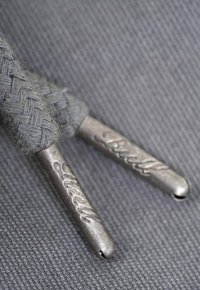 Metal aglets attached to textured grey braided laces, featuring engraved detailing. Background is a smooth grey fabric.