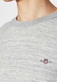 Pull en tricot gris avec un motif texturé et un col rond. Présente un petit accent de logo sur la poitrine en rouge, blanc et bleu.