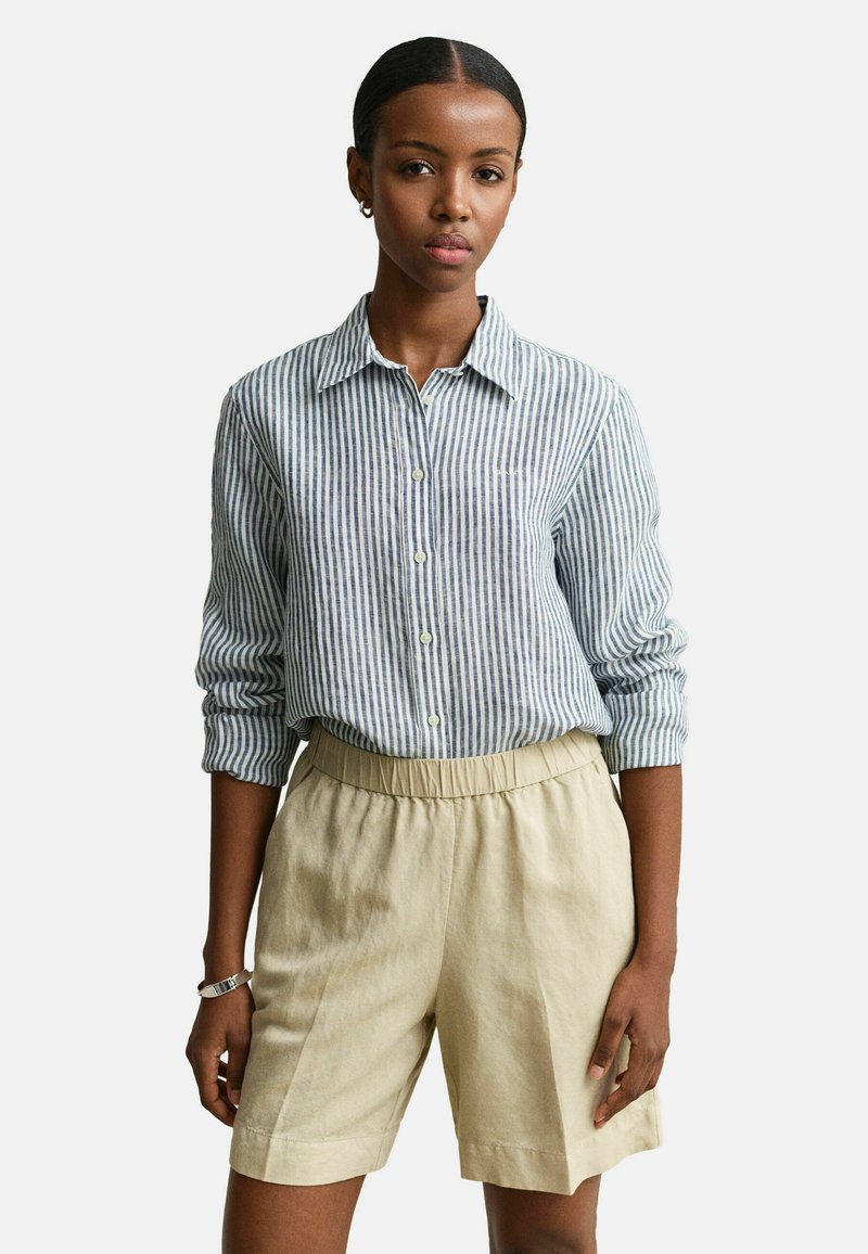 Vrouw draagt een blauw-wit gestreept overhemd met verticale strepen en opgerolde mouwen, beige shorts met elastische tailleband en een zilveren armband.