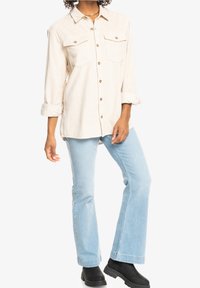 Beige kordfløjl skjorte med knapper og to brystlommer. Sammen med lyseblå flagrende jeans og sorte ankelstøvler. Langærmede ærmer er opsmøgede.
