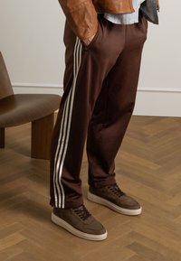 Brown athletic pants with white side stripes, paired with brown sneakers featuring a textured fabric and a white sole.