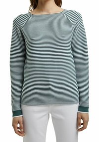 Femme portant un haut à manches longues rayé horizontalement vert et blanc, associé à un pantalon blanc, debout les bras détendus le long du corps.