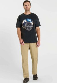 Zwart T-shirt met korte mouwen, bedrukt met een afbeelding van een helm en tekst, gecombineerd met kaki broek en donker schoeisel. Eenvoudig, casual ontwerp.