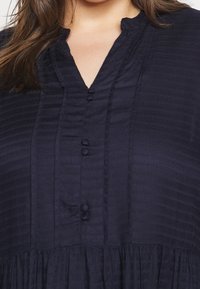 Blusa azul marino con un patrón de rayas texturizado, que presenta una tapeta con botones, un pequeño cuello y una sección fruncida en el dobladillo.