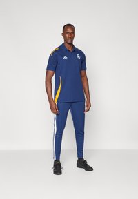Sada námořnických modrých sportovních polo triček a kalhot z lehkého materiálu, se žlutými akcenty a bílými bočními pruhy, s logem na hrudi.
