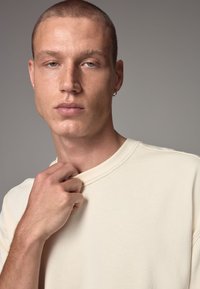Beige cotton short-sleeve T-shirt with a rounded neckline and visible stitching details. Model's hand tugs at collar, showing texture.