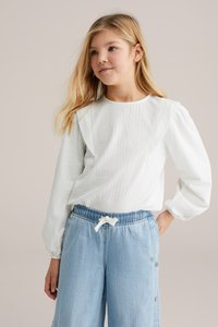 Jeune fille aux longs cheveux blonds portant un haut blanc texturé à manches longues et un pantalon large en denim bleu clair.
