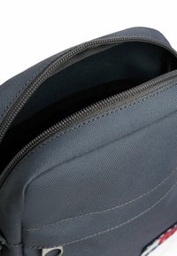 Grå stoftaske med lynlås i toppen, der har struktureret materiale og en glat inderside. Inkluderer en frontlomme med syningsdetaljer.