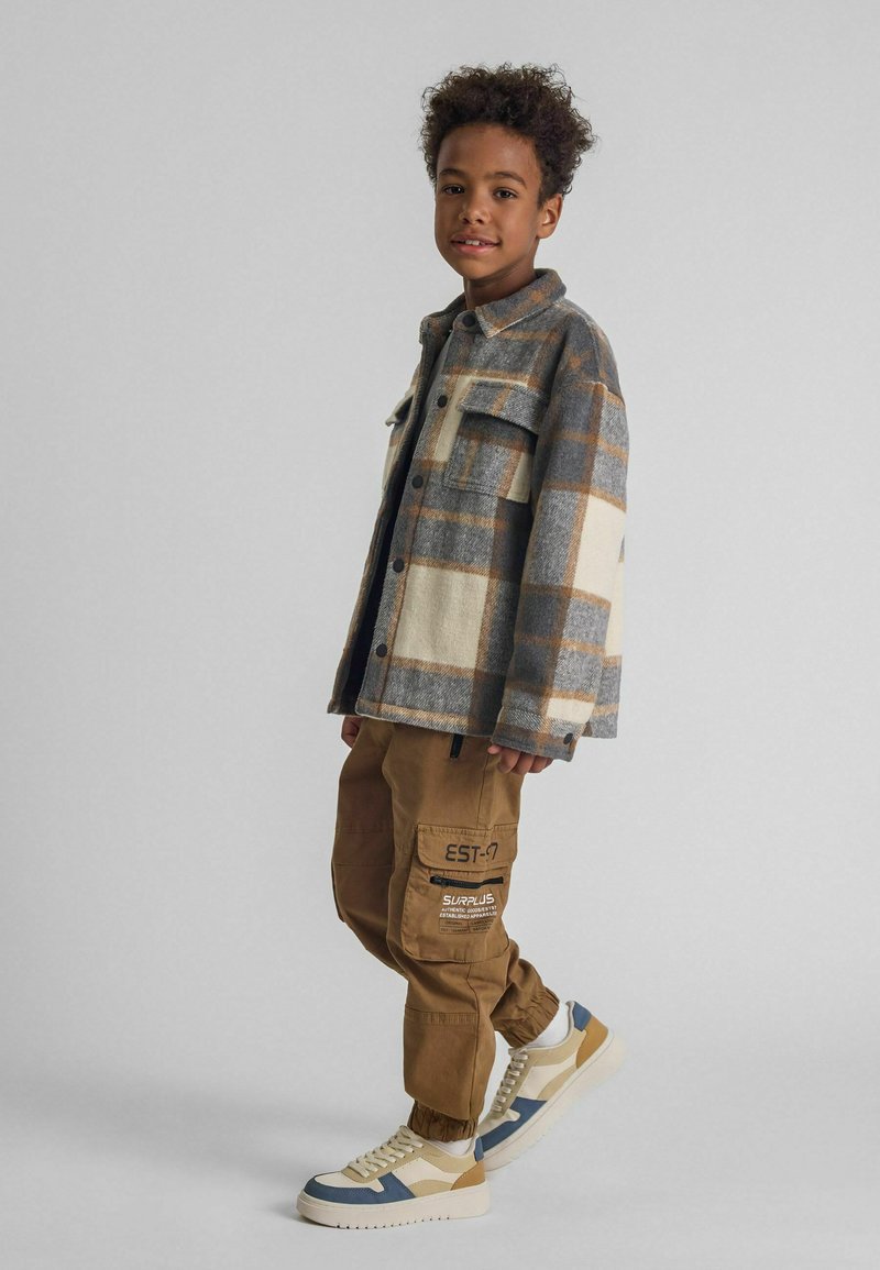 Flanelljacke mit grauem und cremefarbenem Karomuster, braune Cargo-Hosen und weiße Sneakers mit blauen und beige Akzenten. Lässiger Streetwear-Stil.