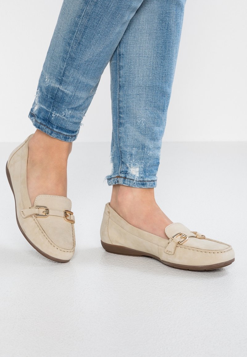 Pieds portant des mocassins beiges avec des accessoires dorés, associés à un jean denim bleu clair légèrement usé sur un fond blanc uni.